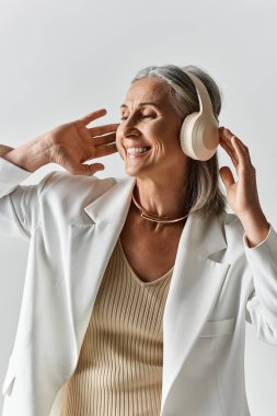 Gri saçlı zarif bir kadın, kulaklık ve beyaz bir takım elbise takıp gülümsüyor ve anın tadını çıkarıyor..