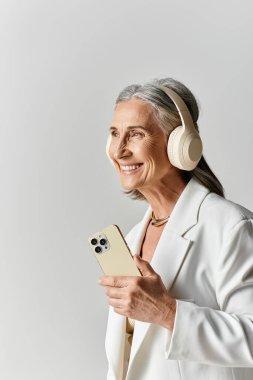 Gri saçlı zarif olgun bir kadın kulaklıkla müzik dinlerken şık beyaz bir takım giyer..