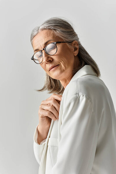 Красивая зрелая женщина с седыми волосами изящно позирует в шикарном белом костюме на приглушенном фоне.