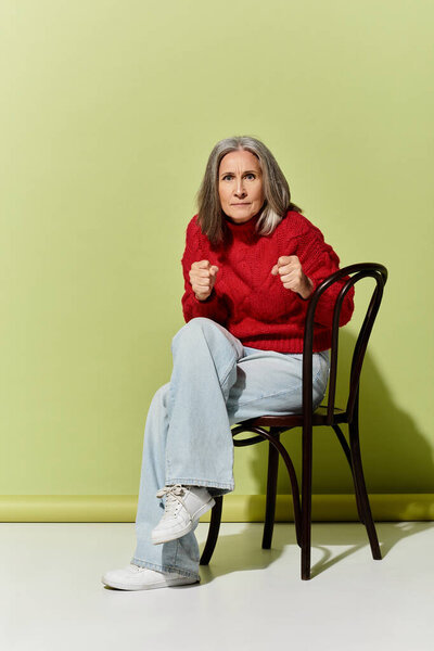 Стильная зрелая женщина без усилий источает уверенность в красном свитере, сидящем на стуле, демонстрируя зимнюю моду.