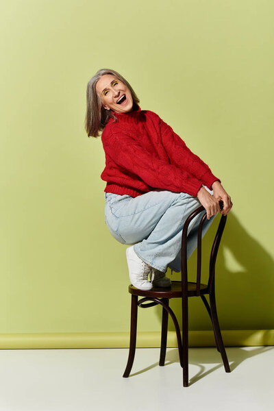 Веселая зрелая женщина демонстрирует свой стильный зимний наряд, игриво сидя на стуле в уютной обстановке.