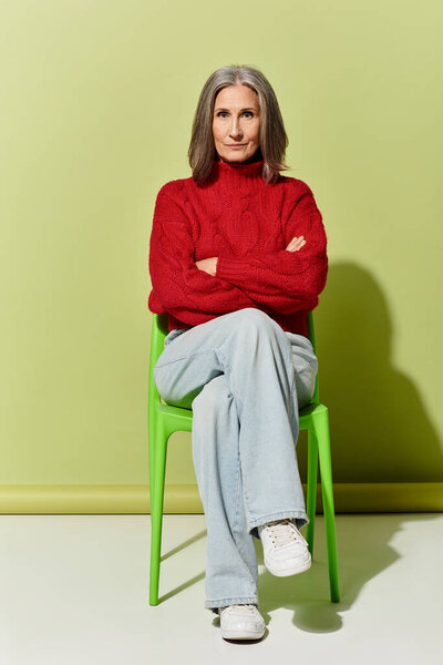 Стильная зрелая женщина с седыми волосами источает уверенность в красном свитере, сидящем на зеленом стуле.