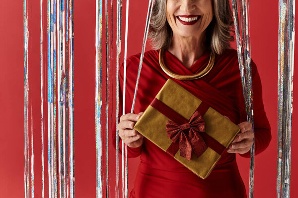 A beautiful mature woman in red attire joyfully holds a gift, celebrating Christmas and the New Year with delight.