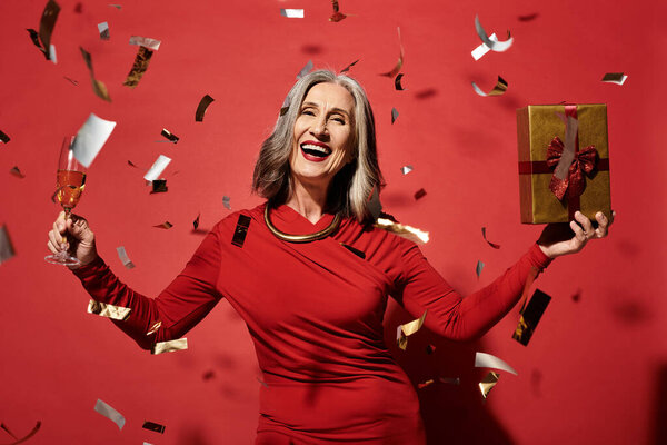 A cheerful mature woman with grey hair enjoys Christmas and New Year, holding a gift and a glass of champagne.