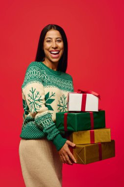 A cheerful woman in a festive sweater holds beautifully wrapped gifts, ready for celebration. clipart