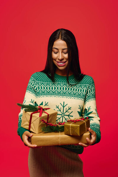 Молодая женщина ярко улыбается, держа в руках завернутые подарки на праздничном красном фоне.