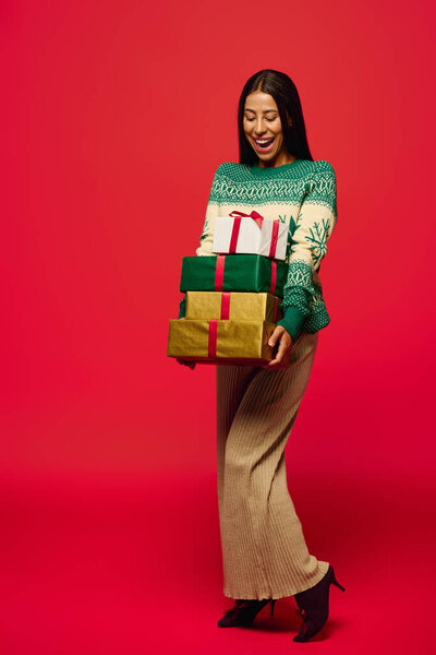 Молодая красивая женщина улыбается, держа в руках несколько упакованных подарков на ярком красном фоне.
