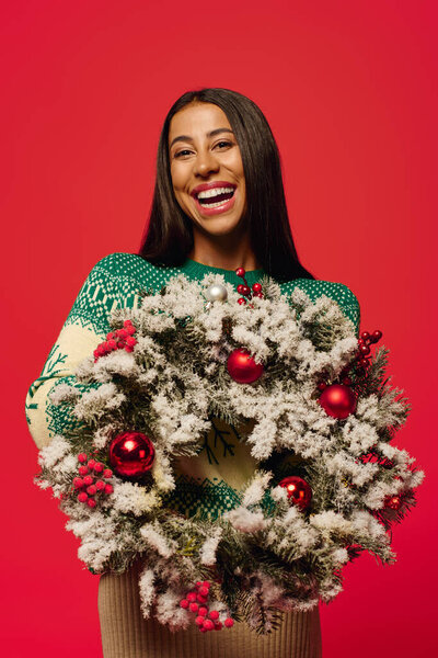 A smiling woman showcases a beautifully decorated Christmas wreath with holiday cheer.