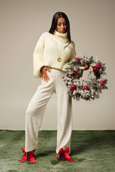 The young woman elegantly poses with a decorative wreath and showcases her festive outfit.