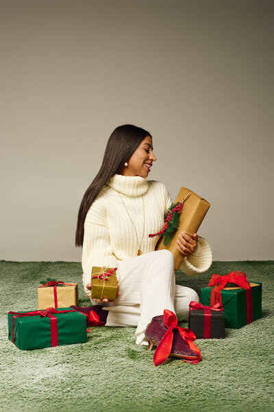 Молодая женщина сияет от радости, когда она держит праздничные подарки на мягком зеленом ковре.