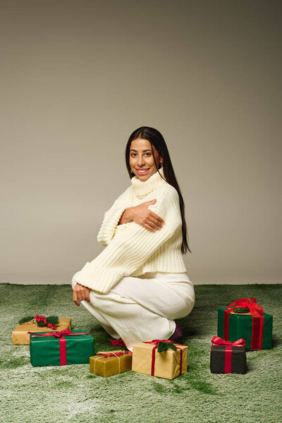 Ошеломляющая молодая женщина в уютном наряде радостно преклоняет колени рядом с красочными обернутыми подарками на мягкой траве.