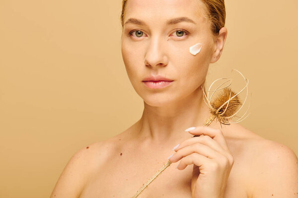 Молодая красивая женщина плюс размер мягко наносит крем на лицо, держа сушеный цветок.