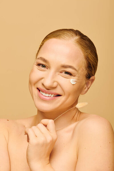 Радостный плюс размер женщина изящно наносит крем на лицо, улыбаясь в камеру.