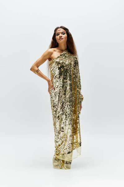 Элегантная богиня в блестящем золотом платье изящно позирует, излучая уверенность в безмятежном пространстве студии.