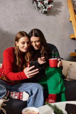 Renkli kazaklı kız arkadaşlar, neşeli sezonu birlikte kutlarken kahkaha ve neşeyi paylaşıyorlar, akıllı telefona bakıyorlar.