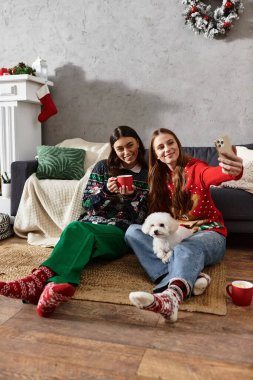 Samimi kazaklar giyen iki kız arkadaş şenlikli içeceklerin ve şirin Bichon frizleriyle selfie çekmenin keyfini çıkarıyorlar..