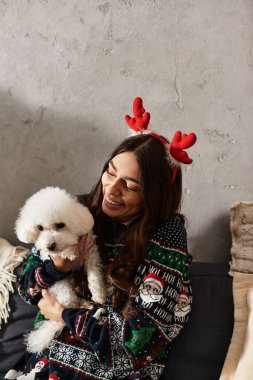 A young woman in a festive sweater joyfully celebrates the holidays with her cute Bichon frise at home. clipart