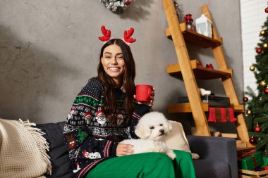 Şenlikli giyinmiş genç bir kadın, neşeli Noel dekorunun yanında sevimli Bichon Frise 'siyle tatilin tadını çıkarıyor..