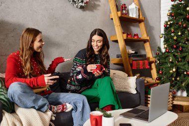 Two friends celebrate the holiday season together, sipping drinks in their cozy sweaters at home. clipart