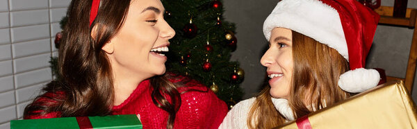 Девушки разделяют радость и смех вместе, разворачивая подарки под елкой в помещении.