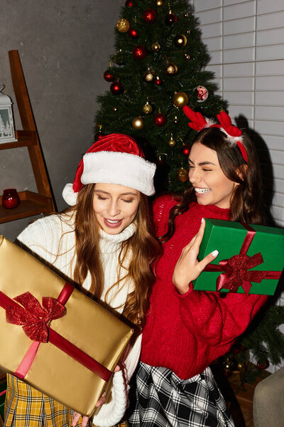 Две девушки с волнением обмениваются подарками возле праздничной елки, обнимая праздничное настроение и тепло.