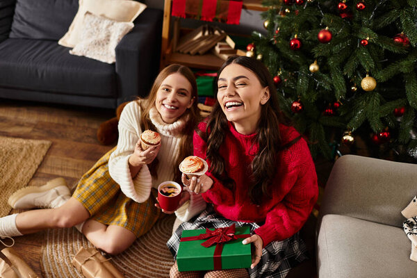 Две девушки радостно наслаждаются праздничными угощениями и подарками у елки, празднуя вместе сезон.