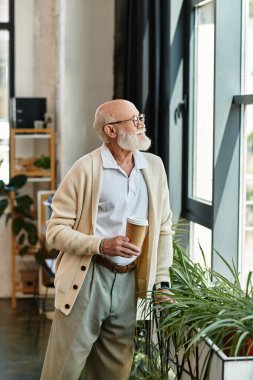 İyi bakımlı yaşlı bir adam, şık bir çalışma alanında kahve fincanı tutarken düşünceli bir şekilde yansıtır..