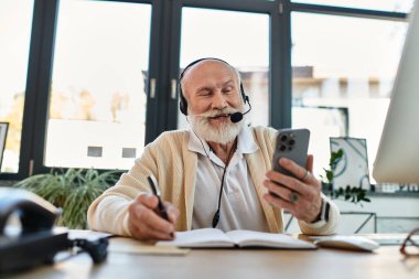 Sakallı kıdemli bir işadamı telefonunu kullanırken gülümsüyor. Modern bir çalışma alanında zahmetsizce birden fazla iş yapıyor..