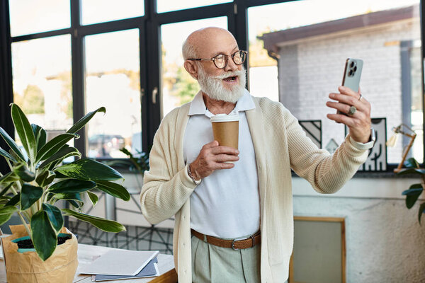 Старший бизнесмен с бородой счастливо взаимодействует по видеосвязи, держа в руках чашку кофе..