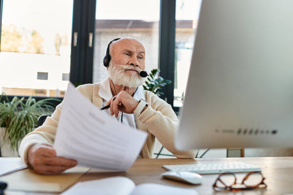 A well-dressed senior businessman reviews documents while using a headset in a contemporary office setting.