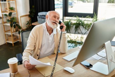 Modern ofis masasında evrak işleriyle uğraşan şık bir kıdemli işadamı telefonda konuşuyor..
