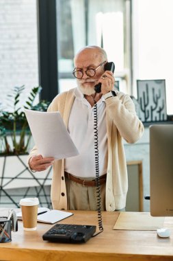 İyi giyimli bir üst düzey yönetici, çağdaş bir ofiste telefonla konuşurken belgeleri gözden geçirir..