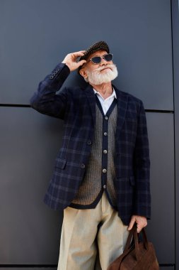 Keskin giyimli, sakallı son sınıf öğrencisi, güz mevsimini kucaklarken çağdaş bir yapının yanında duruyor..