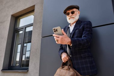 Şık bir son sınıf öğrencisi şık bir binanın yanında duruyor, çantasını tutarken telefonunu kontrol ediyor..