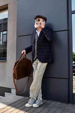 Damalı ceketli ve düz şapkalı şık bir son sınıf öğrencisi modern bir binanın önünde bir telefon görüşmesi yapıyor..