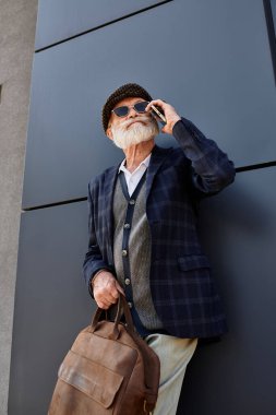 A fashionable senior man talks on his phone, standing near a modern building on a clear autumn day. clipart