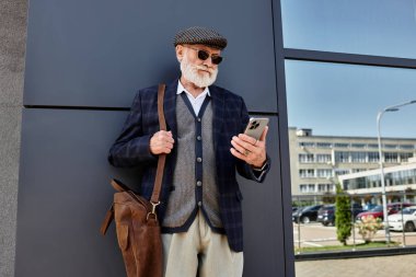 İyi giyimli bir son sınıf öğrencisi, sonbaharda akıllı telefonuna takılı kalmış çağdaş bir yapının yanında gelişigüzel durur..