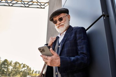 Sakallı, modaya uygun, üst düzey bir adam çağdaş bir yapının yanında durup telefonunu kontrol ediyor..