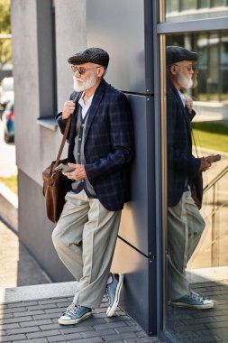 Sakallı yaşlı bir adam modern bir binanın yanında rahatlar. Sonbahar havasıyla kolayca harmanlanır..