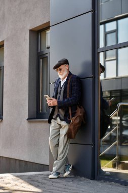 İyi giyimli yaşlı bir beyefendi, sonbahar havasının tadını çıkarırken telefonunu kontrol ederek bir binaya yaslanıyor..
