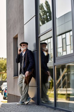 Sakallı şık bir kıdemli adam, sonbaharda çağdaş bir binanın yanında telefonuyla sohbet ediyor..