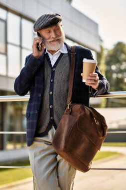 İyi giyimli, son sınıf öğrencisi, sakallı bir adam telefonda, çağdaş bir binanın yanında kahve içiyor..