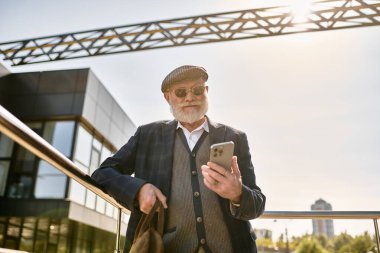 Üst düzey şık ceketli ve düz şapkalı şık bir adam telefonunu çağdaş bir binanın dışında kontrol ediyor..