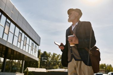 Sakallı şık bir son sınıf öğrencisi, modern bir binanın sonbahar manzarasına bakarken elinde kahve fincanı tutuyor..