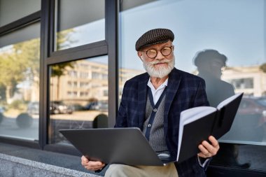 Sonbahar renklerinin ortasında çağdaş bir binanın yanında dizüstü bilgisayarı olan şık bir kıdemli adam oturuyor..