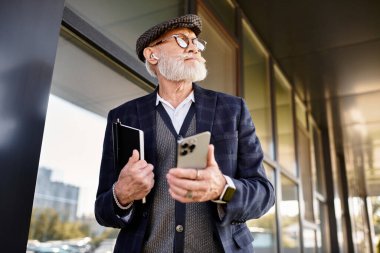 Modern bir binanın önünde duran şık, yaşlı bir adam elinde telefonuyla sonbahar havasının tadını çıkarıyor..