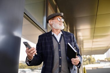 Sakallı şık bir son sınıf öğrencisi elinde bir telefon ve defter tutuyor ve modern mimarinin sonbahar havasını yansıtıyor..