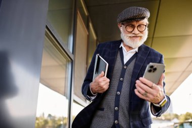 Sakallı şık bir son sınıf öğrencisi, sonbaharda çağdaş bir binanın yanında dururken akıllı telefonunu kontrol ediyor..