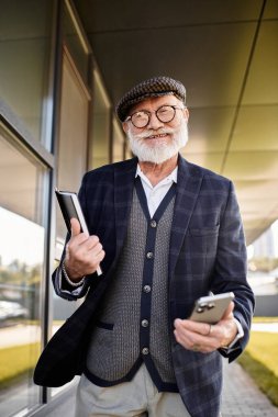 Sakallı son sınıf öğrencisi elinde tablet ve telefonla çağdaş bir yapının yanında sonbahar tarzını sergiliyor..