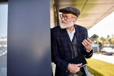 Seçkin bir kıdemli beyefendi modern bir binanın yanında duruyor, telefonuna bakarken güneşli bir sonbahar gününün tadını çıkarıyor..